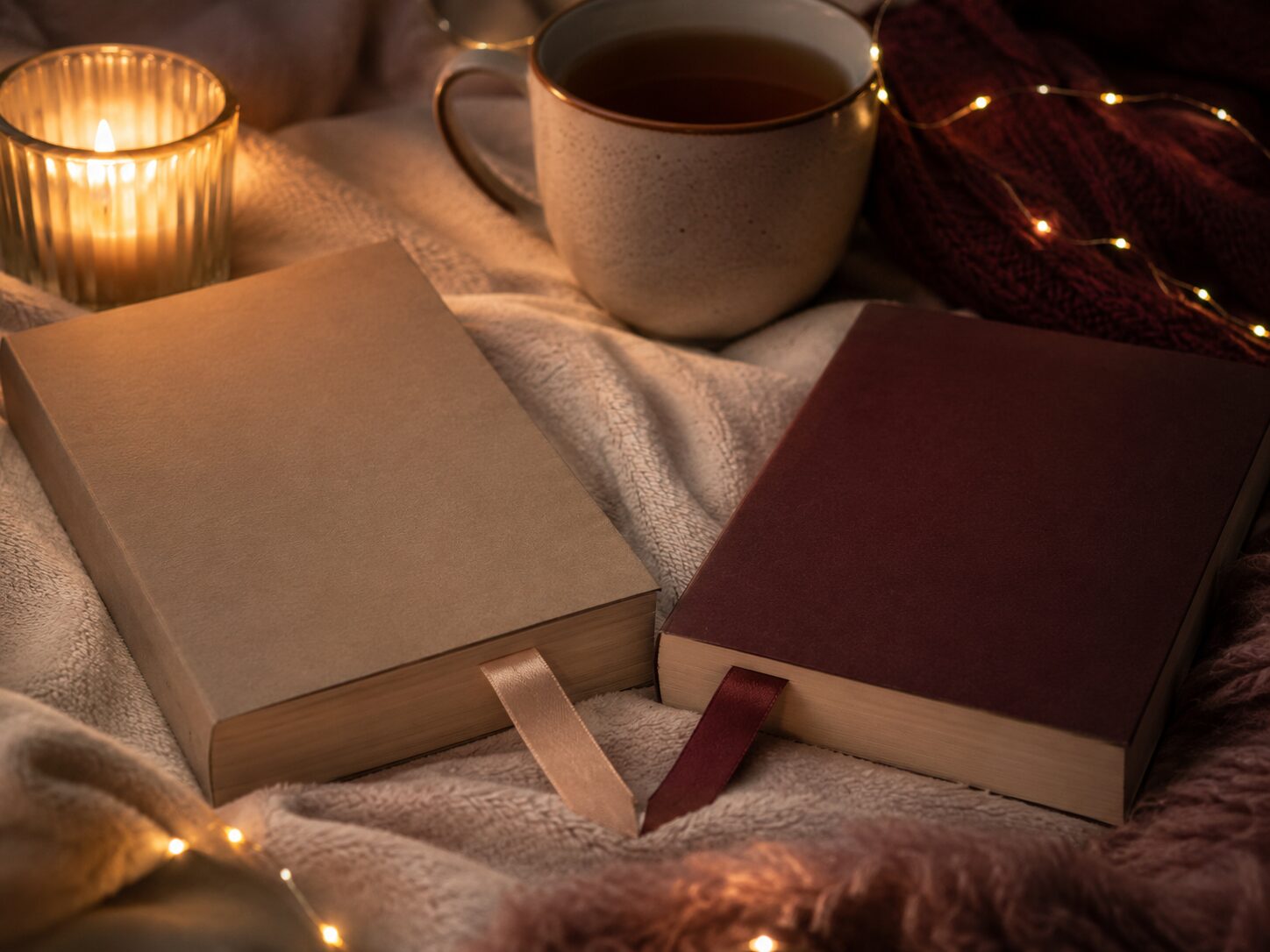 Two romance books with ribbon bookmarks beside a candle and tea mug with fairy lights on a cosy blanket