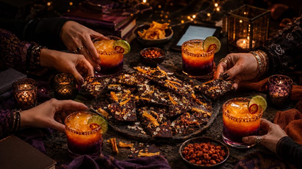 Hands reaching across a candlelit table for Dragon's Fire Margaritas with chilli-salt rims, around a slate board of Dragon Scale Bark scattered with dried mango and toasted coconut.